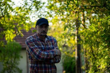 Kendinden emin, kareli gömlekli, kollarını kavuşturup köydeki kameraya bakan olgun bir adam..