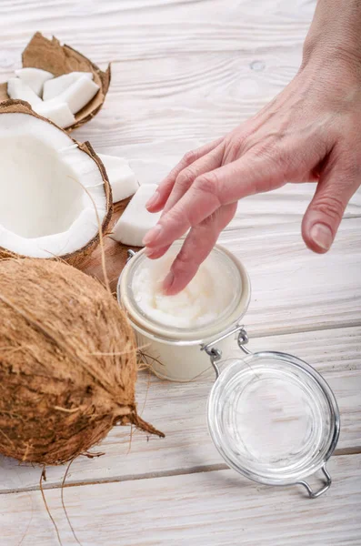 Coconut oil on human palm and in airtight glass jar with shell pieces aside on white wooden table