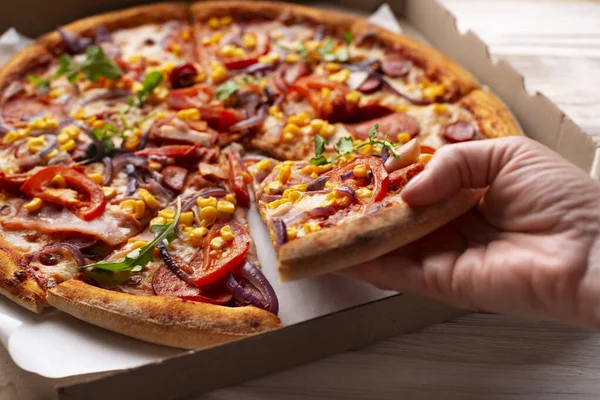 Human caucasian hand takes slice of large pizza from carton box on white wooden table