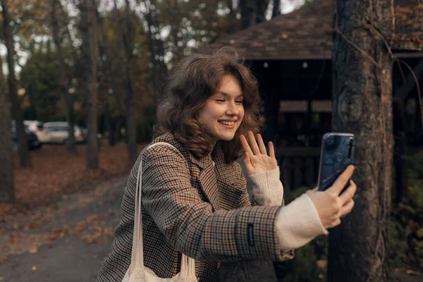 年轻的微笑女孩在秋天的公园里散步 用智能手机自拍自拍 开心的心情时尚潮流 — 图库照片