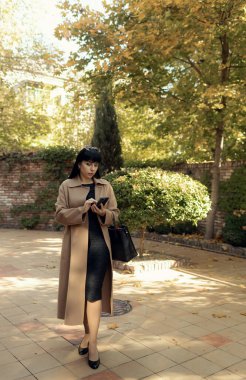 Orta yaşlı bir iş kadını akıllı telefondan konuşuyor. Havalı bir düşüşle dışarı çıkıyor. Kadın stili model şehir caddesinde yürüyor. Sonbahar trendi, moda kıyafeti