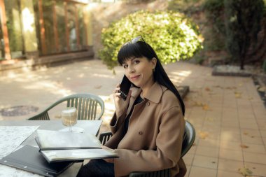 Orta yaşlı bir iş kadını akıllı telefondan konuşuyor. Havalı bir düşüşle dışarı çıkıyor. Kadın stili model şehir caddesinde yürüyor. Sonbahar trendi, moda kıyafeti