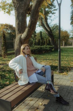 Parkta yüzünde mutlu bir gülümseme olan kadın, seviniyor. Şehir parkının bulanık arka planında yarı sezon kıyafetleri giymiş bir kız.