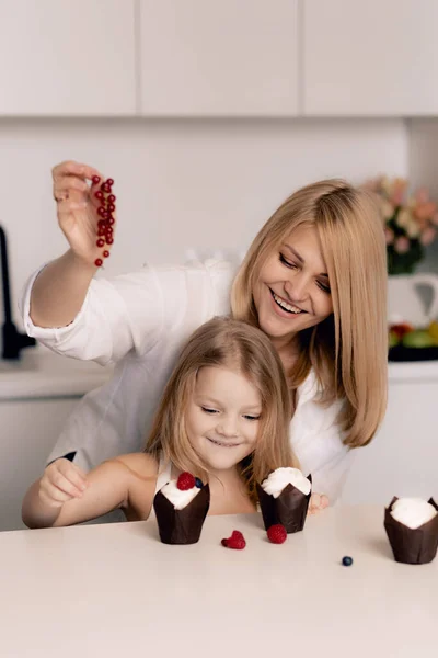 Pastry chef confectioner young caucasian woman with her daughter decorate a cake and cupcakes in the kitchen.