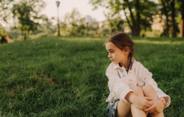 Yeşil bir ormanda temiz hava soluyan rahat küçük kız. Güneşli yaz gününde parkta yeşil çimlerin üzerinde oturan beyaz elbiseli güzel bir genç kız. Büyüleyici Ukrayna çocuk portresi. Mutlu bir çocukluk ve açık hava eğlencesi konsepti.