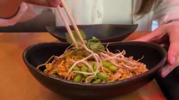 ストリートフード 皿の上にはタイ麺がたくさんある 女性のクローズアップは タイのカフェでプレートからアジアの麺を取るために箸を使用しています — ストック動画