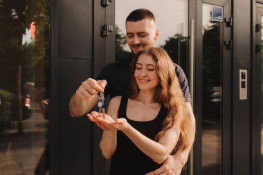 Heyecanlı genç eşlerin ev sahiplerinin tuttuğu anahtarlara odaklan. Mutlu evli çift yeni eve taşınmayı kutluyor, anahtarları gösteriyor, apartmanda duruyor, gayrimenkul ipoteği konsepti....