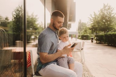 Genç bir adam iş için cep telefonu kullanıyor ve oğlunun elini kafede tutuyor. Baba olmak ve her yerde çalışmak. Serbest ve ağ oluşturma kavramı.