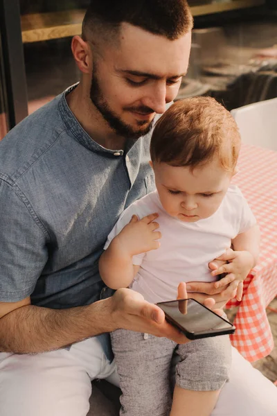 Genç bir adam iş için cep telefonu kullanıyor ve oğlunun elini kafede tutuyor. Baba olmak ve her yerde çalışmak. Serbest ve ağ oluşturma kavramı.