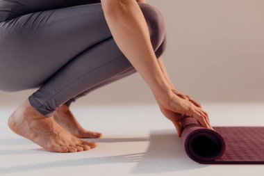 Genç bir kadın, stüdyoda yoga dersinden önce ya da sonra paspas çeviriyor. Hafif bir arka planda otururken yoga yapan bir kadının yakın plan bacakları..