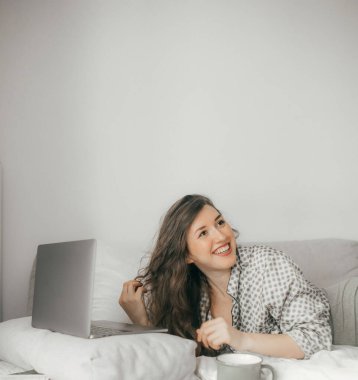 Yatak odasında poz veren genç bir kadın, makyajı temiz, dizüstü bilgisayarı evde oturuyor ve mesaj yazıyor. Yumuşak odaklı ve gürültülü fotoğraf konsepti. Teknoloji ve içerik pazarlama kavramı.