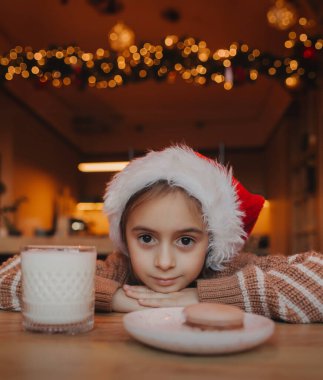 Noel Baba şapkalı şirin bir kız ve evde lezzetli bir Noel kurabiyesi ve süt var. Xmas evde tatil kutlaması.