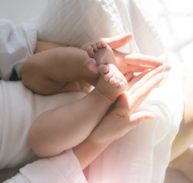 Anne yeni doğmuş bebeğin çıplak topuklarını tutuyor. Aile ve ev kavramı. Sağlık, pediatri. Evde sıcak bir sabah. Annelik. Güneşli fotoğraflar, yumuşak odak, aşırı pozlama..