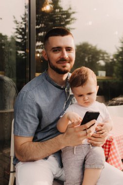 Genç bir adam iş için cep telefonu kullanıyor ve oğlunun elini kafede tutuyor. Baba olmak ve her yerde çalışmak. Serbest ve ağ oluşturma kavramı.
