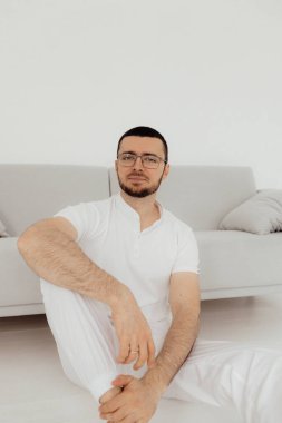 Ciddi, sakallı, erkek terapist. Tıp üniformalı. Kollarını kavuşturup, beyaz arka planda, ışık kliniğinde kameraya bakıyor..