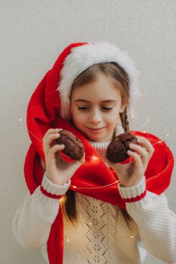 Kazaklı ve Noel Baba şapkalı tatlı bir genç kız bir ışık duvarının arkasında çikolatalı kurabiye tutuyor ve yiyor. Şenlikli Yeni Yıl yemeği konsepti.