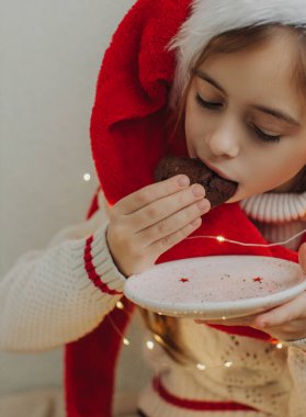 Kazaklı ve Noel Baba şapkalı tatlı bir genç kız bir ışık duvarının arkasında çikolatalı kurabiye tutuyor ve yiyor. Şenlikli Yeni Yıl yemeği konsepti.