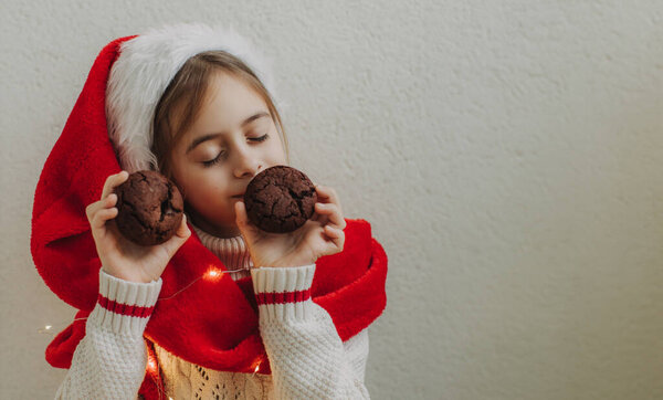A cute teenage girl in a pullover and Santa Claus hat holds and eats a chocolate cookie against the background of a light wall. Festive New Year food concept.