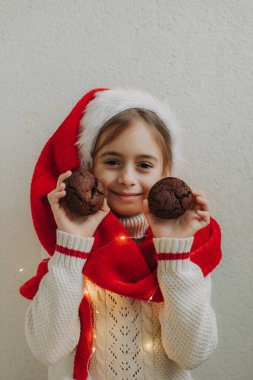 Kazaklı ve Noel Baba şapkalı tatlı bir genç kız bir ışık duvarının arkasında çikolatalı kurabiye tutuyor ve yiyor. Şenlikli Yeni Yıl yemeği konsepti.