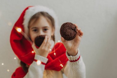 Kazaklı ve Noel Baba şapkalı tatlı bir genç kız bir ışık duvarının arkasında çikolatalı kurabiye tutuyor ve yiyor. Şenlikli Yeni Yıl yemeği konsepti.