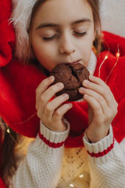 Kazaklı ve Noel Baba şapkalı tatlı bir genç kız bir ışık duvarının arkasında çikolatalı kurabiye tutuyor ve yiyor. Şenlikli Yeni Yıl yemeği konsepti.