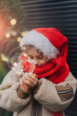 Noel Baba şapkalı sevimli genç kız LED çelenkle Noel ağacını süslüyor ve dışarıda bir dilek tutuyor. Çocuklarla mevsimsel kış tatillerini kutlarız..