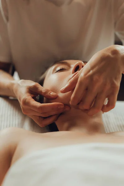 Hands of female chiropractor massaging face of young woman lying on massage table. Visceral massage. Concept of physical therapy treatment, neck pressure point.