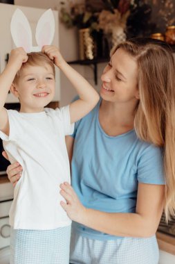Paskalya için hazırlanıyorum. Aile evde. Paskalya Ailesi gelenekleri. Sevgi dolu bir anne ve kartondan tavşan kulakları olan mutlu küçük bir çocuk. Evdeki rahat açık mutfakta gülümsüyor..