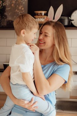 Paskalya için hazırlanıyorum. Aile evde. Paskalya Ailesi gelenekleri. Sevgi dolu bir anne ve kartondan tavşan kulakları olan mutlu küçük bir çocuk. Evdeki rahat açık mutfakta gülümsüyor..