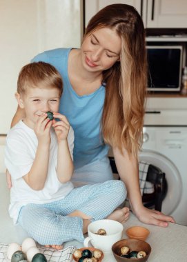 Paskalya için hazırlanıyorum. Aile evde. Paskalya Ailesi gelenekleri. Sevgi dolu bir anne ve kucağında küçük mutlu bir çocuk. Boyalı çok renkli Paskalya yumurtaları. Evdeki rahat açık mutfakta gülümsüyor..