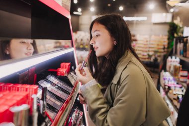 Güzeller güzeli gençlik testleri ve kozmetik ürünleri bir güzellik mağazasından almak. Kozmetik mağazasında ruj deneyen genç bir kız. Perakende ve tüketim. Kişisel bakım ve modern kozmetoloji. Yan görünüm.