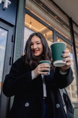 Pencerenin yanındaki kafede elinde kağıt bardakta iki kahve olan sevimli Koreli kızın portresi. Genç kız kapuçino içiyor ve kameraya bakıyor..