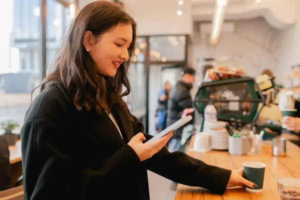 Pencerenin yanındaki kafede akıllı telefona bakıp kahve içen sevimli Koreli kız portresi. Kapuçino içen ve cep telefonuyla konuşan bir kadın..