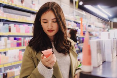 Genç kız perakende mağazasında elinde bir şişe krem tutuyor. Güzeller güzeli bir kadın güzellik salonundan kozmetik ürünleri alıyor. Yan görüş. Kişisel bakım ve modern kozmetoloji.