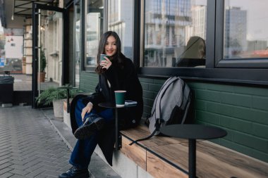 Pencerenin yanındaki kafede elinde kağıt bardakta iki kahve olan sevimli Koreli kızın portresi. Genç kız kapuçino içiyor ve kameraya bakıyor..