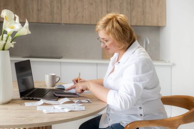 Üst düzey iş kadını elinde dizüstü bilgisayarla kağıt para tutuyor. Yaşlı kadın banka hesaplarını yönetiyor, bütçe vergilerini hesaplıyor, banka borçları için emeklilik ödemelerini planlıyor mutfak masasında oturuyor..