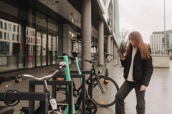 スマートフォンを使用して電動スクーターや自転車を借りる学校の年齢の少女は モバイルアプリを通じて無接触の支払いを行います 秋冬シーズンの市内の自転車レンタル — ストック写真