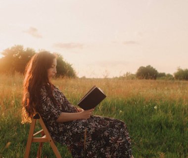 Kitabı olan sakin bir kadın anın tadını çıkarıyor ve doğada yavaş bir yaşam sürüyor. Yeşil turizm kavramı, yavaş yaşam, ekoloji. Doğadaki kitaplar, klasik tondaki renkler ve yumuşak odaklanma. Dizinin bir parçası.
