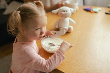 Baby girl eats breakfast and switching channels on the TV with the remote control. Harmful upbringing of a child with gadgets. Children and technology concept. Part of the series.