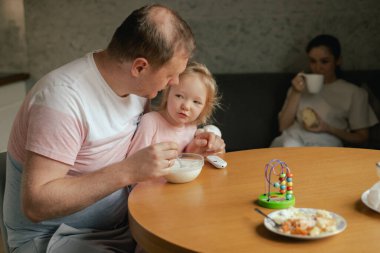 Kızı kollarında onu besleyen bir babanın yakın çekimi. Annem mutfak işleriyle meşgul. Ailede cinsiyet karışımı ve rol değişimi kavramı. Dizinin bir parçası.