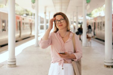 Şehir yolculuğu konsepti. Telefonundaki bilete bakan ve istasyonda ulaşımı bekleyen turist kadın, başvuru formunda internetten bilet ayırtıyor. Uzayı kopyala Dizinin bir parçası.