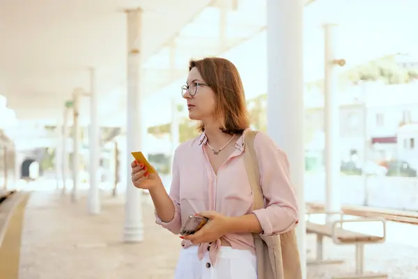 stock image City travel concept. Tourist Woman looking at a ticket on her phone and waiting for transport at station, passenger books a ticket online through in the application. Copy space. Part of the series.