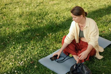 İletişim, sosyal medya ve yaz parkında haber uygulamaları için telefon ve iletişim ağı olan orta yaşlı bir kadın. Yoga minderinde oturan kadın. Doğayla birlik, spor yoga konsepti.