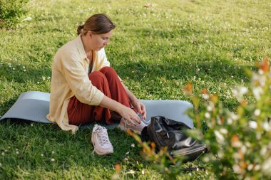 Orta yaşlı bir kadın dışarıda yoga yapmak için ayakkabılarını çıkarıyor. Yoga eğitmeni parkta yoga dersi için hazırlanıyor. Doğa kavramıyla birlik.