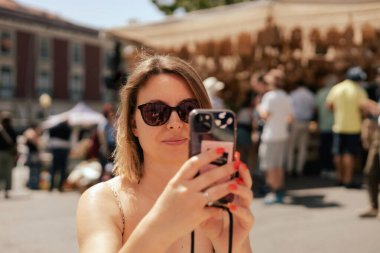 Orta yaşlı bir kadın bit pazarında akıllı bir telefonla fotoğraf çekiyor. Garaj satışı, giysileri tekrar kullanma, ikinci el ve ekolojik tüketici. Yakın plan. Seçici odak.