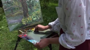 Ressam kız resim çizdikten sonra ellerindeki boyayı ve sehpayı sallar. Genç bayan ressam güneş ışığında yaz parkında resim yapıyor. Sanat ve hobi konsepti.