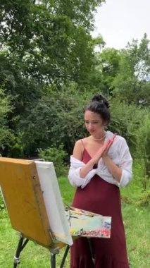 Ressam kız, resim çizdikten sonra elindeki ve sehpadaki boyayı silkeliyor. Genç bayan ressam güneş ışığında yaz parkında resim yapıyor. Sanat ve hobi konsepti. Bir serinin parçası.