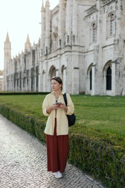 Middle aged traveling woman looks for the way using the online map on her phone and passes the Monteiro dos Jeronimo's in Belem, Lisbon, Portugal. Travel and solo tourism concept. Part of the series. clipart