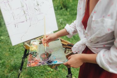 Bir kadının ellerini fırça ve boyalarla etekli bir tuvalin yanına yaklaştır. Farkındalık, sanat terapisi, yaratıcılık kavramı. Doğada plein hava boyama işlemi. Güzel sanatlar, eskiz, eskiz defteri..