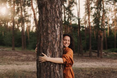 Açık hava eğlence parkında mutlu yetişkin kadın ağaç gövdesine sarılıyor. Çevre ve çevre bakımı kavramı. İnsanlar Dünya gezegenini sever ve ormanların yok oluşunu durdurur.